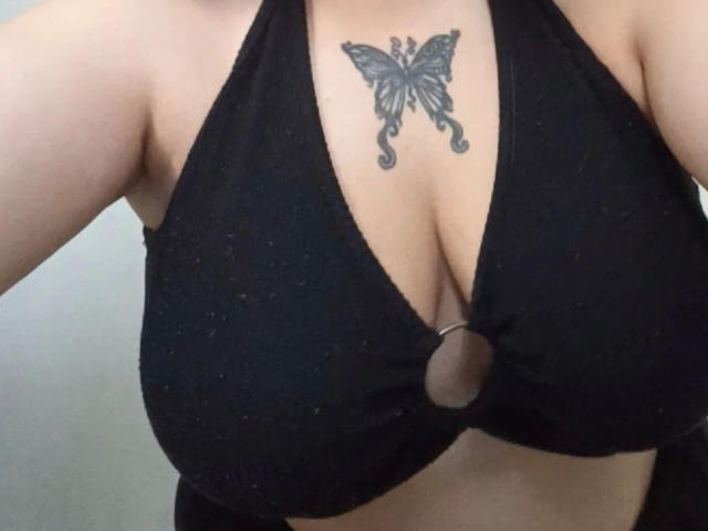 Het zwarte haltertopje van LunaGeil-hot met een ring in het midden, dat haar grote, ronde borsten laat zien en een gedetailleerde vlindertatoeage net boven haar decolleté..