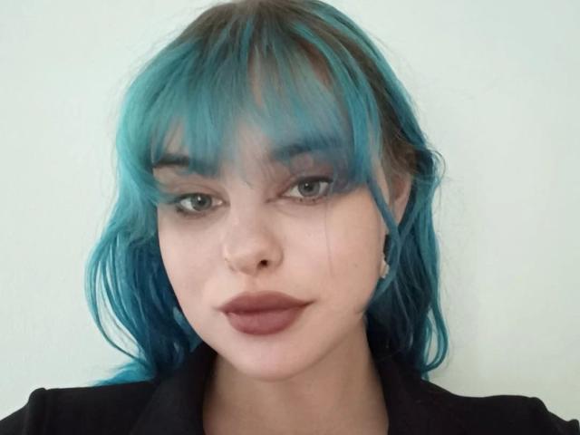 LunaGeil-hot has teal blue hair, full lips with dark pink lipstick, and light green eyes. She's wearing a black top, and her ears have small silver earrings.