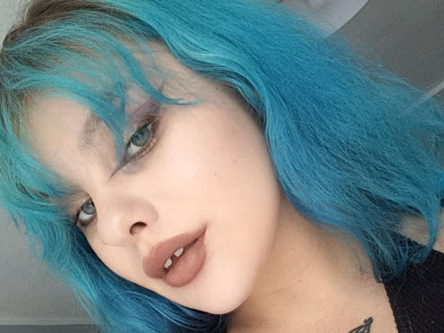 LunaGeil-hot with bright blue hair, pouty brown lips, and a tattoo on her neck, looking directly at the camera with half-closed green eyes.