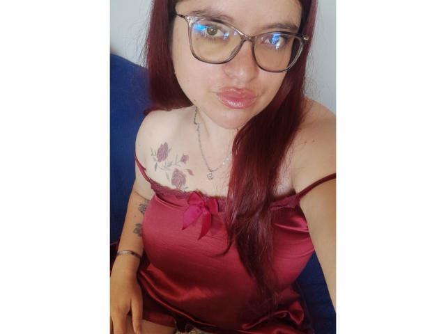 ElizabethFire with red hair, glasses, and a red satin camisole, showing her tits with a small bow and tattoo on her right shoulder, pouting her glossy lips.