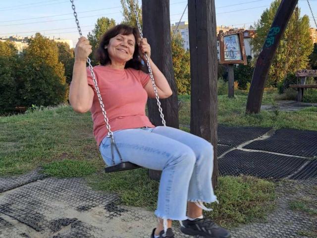IngridWels está sentada em um balanço, segurando as correntes com as duas mãos, sorrindo, vestindo uma camisa rosa, jeans azuis e sapatos pretos. Ela tem seios médios e cabelo castanho escuro..