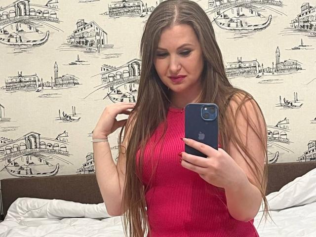HillarySweet with long brown hair, pink dress, holding iPhone, taking selfie, showing her tits, pink lipstick, white bed, brown headboard, patterned wallpaper, holding phone with right hand.