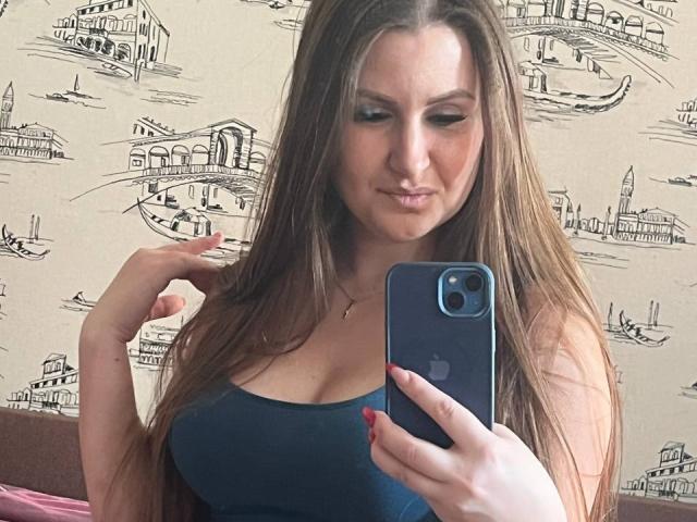 HillarySweet with long brown hair, wearing a blue tank top, showing her big boobs in a mirror selfie, holding a blue iPhone, with red nails, looking down.