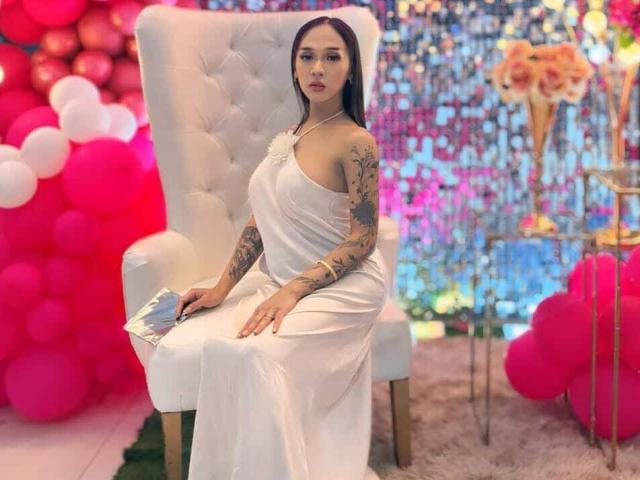 CumPrivateTS with tattoos, sitting on a white tufted chair, wearing a shiny white dress, holding a card, has large tits, long dark hair, pink and white balloons.