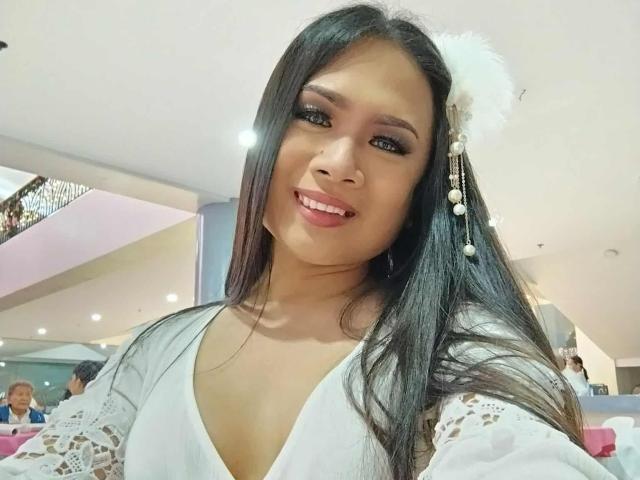 CumPrivateTS has long black hair, brown eyes, and is smiling with pink lipstick. She's wearing a white lace top and a white flower in her hair with pearl earrings.