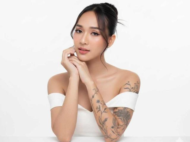 CumPrivateTS with tattoos on her right arm, wearing a white off-shoulder top, hands near her face, dark hair in a bun, soft smile, and a relaxed expression.