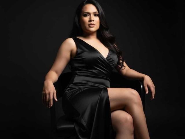 CumPrivateTS is a curvy woman with long black hair, wearing a black satin dress, sitting with her leg crossed, showing her smooth, bare thigh, and looking confident.