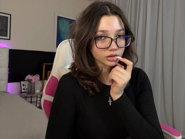 EllieJulse with glasses, brown wavy hair, black long-sleeve shirt, finger in mouth, white and pink chair, cross necklace, looking slightly pouty, in a bedroom.
