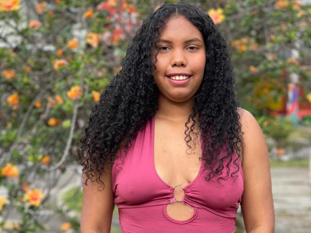 ElizabethMona with curly black hair, medium brown skin, smiling, wearing a pink halter top with a keyhole cutout, showing her small tits, standing outdoors.