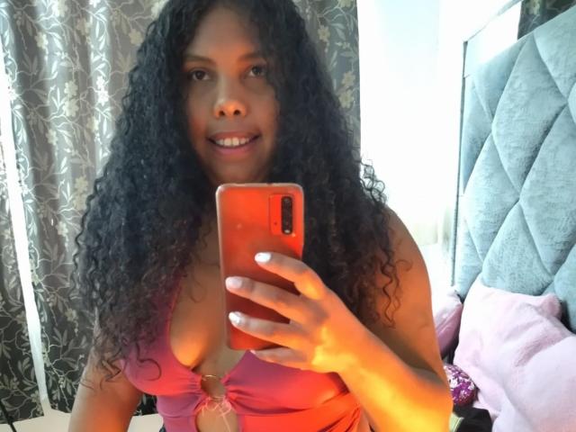 ElizabethMona with curly black hair, wearing a pink top with a keyhole, holding an orange phone, smiling, showing her tits, standing in a bedroom with a gray headboard.