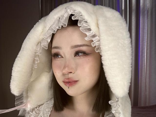 Beauty69Diva with pale skin, dark brown hair, and subtle makeup, wearing a fluffy white hood with frilled edges, has a nose ring and lip piercing, looking to the side.