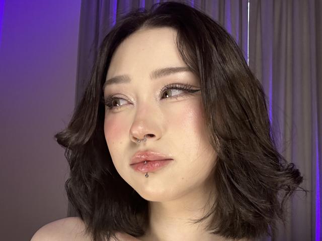 Beauty69Diva has pale skin, dark wavy hair, brown eyes, and a septum piercing with a lip ring. Her pink lips are slightly parted. Soft purple light is in the background.