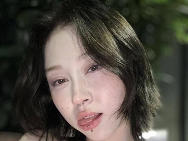 Beauty69Diva with short black hair, nose piercings, lip piercings, light skin, and brown eyes, looking slightly to the side, with a subtle, relaxed expression.