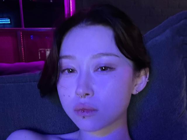 Beauty69Diva with purple-blue lighting, brown hair, nose ring, lip piercing, ear piercing, and slightly parted lips, sitting on a grey couch. Her face is in soft focus.