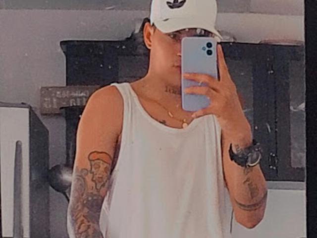 ErosAndree with tattoos on both arms, wearing a white tank top and cap, holding a phone to cover his face, standing in a kitchen, showing his ass.