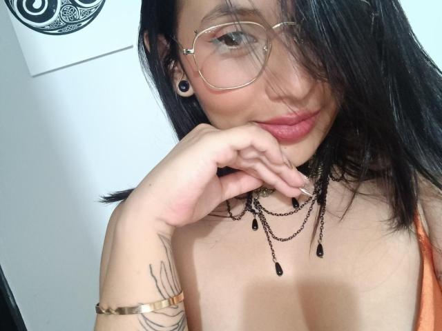 LiliethWolf with black hair, glasses, and a nose ring, smiling, showing off her tits, wearing a necklace, and a gold bracelet, with a tattoo on her right arm.