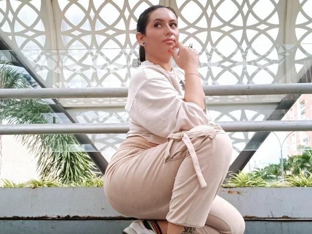 LiliethWolf with dark hair, tan pants, and a white top, squatting, thinking, with her finger on her lips, showing her ass, in a bright, geometric-patterned roofed area.