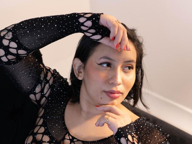 LiliethWolf with medium brown skin, black mesh top showing her tits, red nail polish, ring on finger, black hair, slightly parted lips, looking sideways, hand on head.