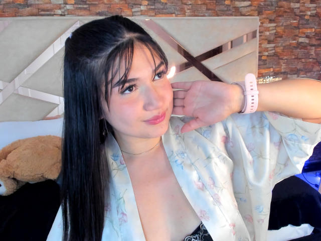 AmyRousse with long black hair, wearing a floral kimono, pink watch, and a black lace bra, smiling softly, hand near cheek, bed with brown teddy in background.
