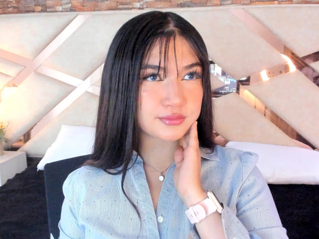 AmyRousse with long black hair, blue eyes, and pink lips, wearing a light blue button-up shirt, pink watch, and necklace, resting her hand on her cheek.