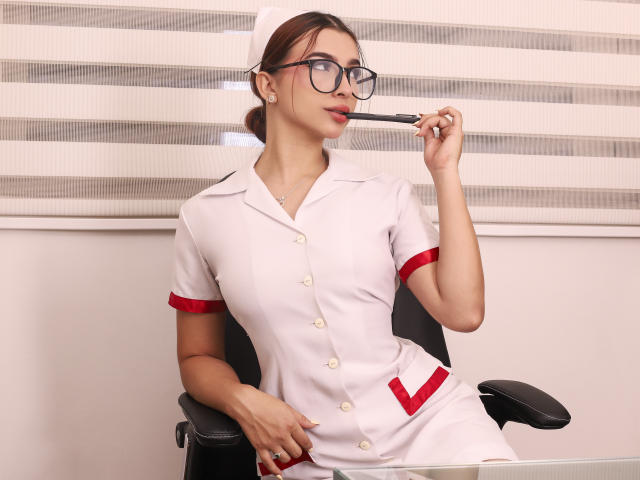 NicolsAdams, wearing a white nurse outfit with red trim, black glasses, and a white cap, sucking on a pen, sitting in a chair, holding a clipboard.