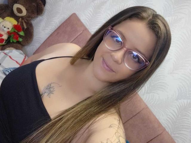 CleareRems has long brown hair, glasses, black top, small tattoo on chest, and a subtle smile, sitting with a pink cushion and brown teddy bear in the background.