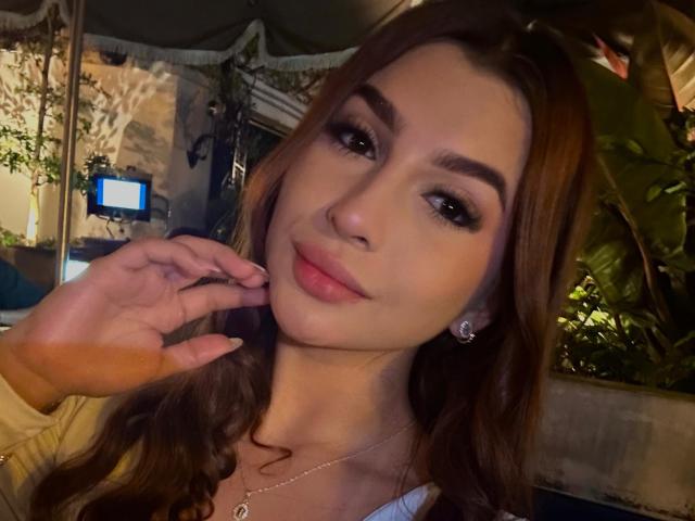 NicolsAdams has brown hair, full eyebrows, dark eyes, pink lips, and is touching her cheek with a hand. She's wearing a necklace and has a subtle smile.