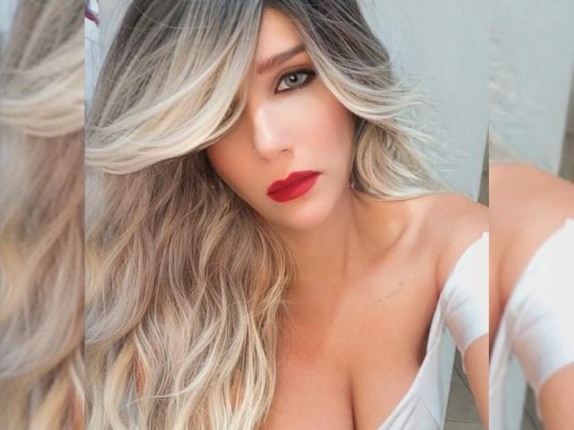 RedVevelt69 with blonde, wavy hair, red lipstick, and a white, off-shoulder top, showing her tits and a reflection of her face in a mirror.