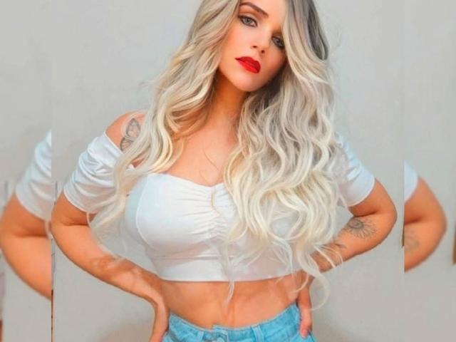 RedVevelt69 has long, wavy blonde hair, red lipstick, white off-shoulder crop top, and blue jeans. She's showing off her tits and tattoos with hands on hips.