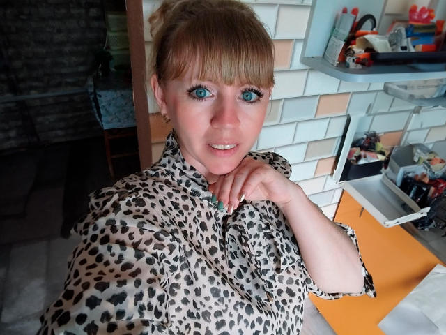 HayleyLaRise with blue eyes, blonde hair, and a leopard print shirt, smiling, her hand near her mouth, wearing a green ring. Her tits are small, she's cute.