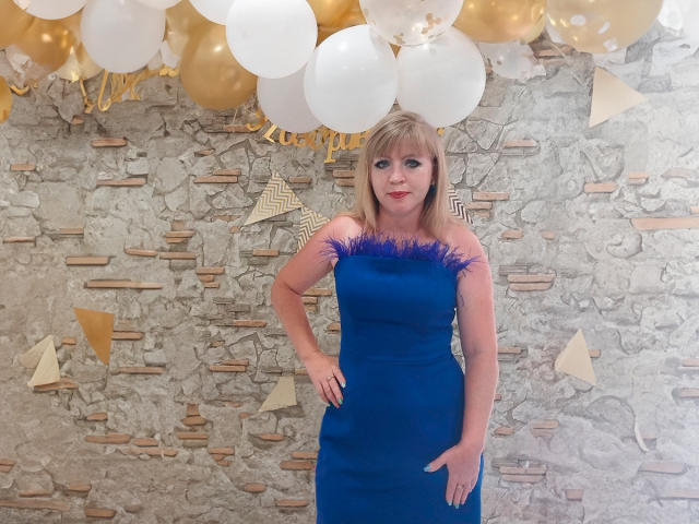 HayleyLaRise has blue dress with purple feather trim, blonde hair, and blue eye makeup, standing with hand on hip, surrounded by white and gold balloons.