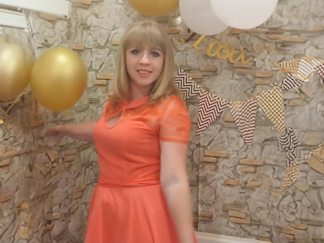 HayleyLaRise with blonde hair, orange dress, and cute smile, holding a gold balloon, standing in front of a stone wall, and white balloons, looking slightly to the left.