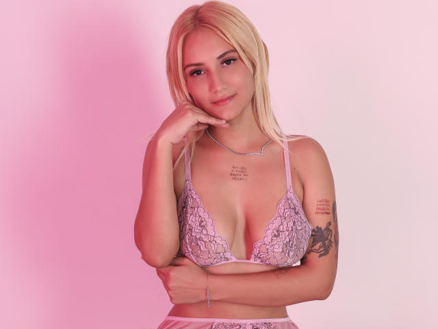 Amber Robb mit blonden Haaren, die ein lila Spitzen-BH trägt, wobei ihre Brüste zu sehen sind, mit Armtätowierungen und einem Armband, sieht süß aus vor einem rosa-weißen Hintergrund..