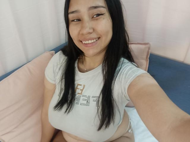 Karollteyes, met lang zwart haar, lachend, draagt een witte crop top die haar grote borsten laat zien, en zit op een roze en blauw bed. Ze houdt een selfie vast..