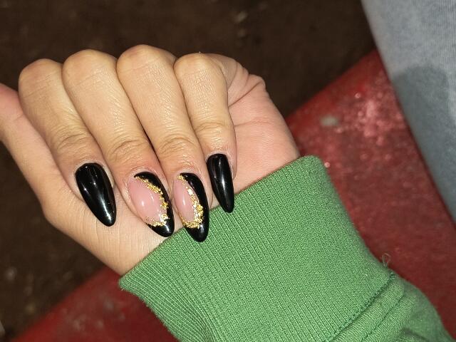 JulietaFoster69's hand with shiny black nails and a gold glitter accent on the ring finger, wearing a green sleeve, against a red and brown background.