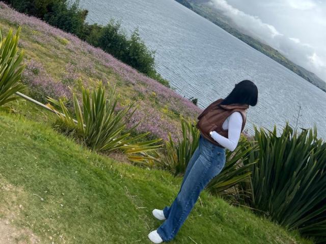 IsabellaJames with black hijab, brown vest, white long-sleeve shirt, blue jeans, and white sneakers, standing on grassy hill with purple flowers and green plants, facing lake.