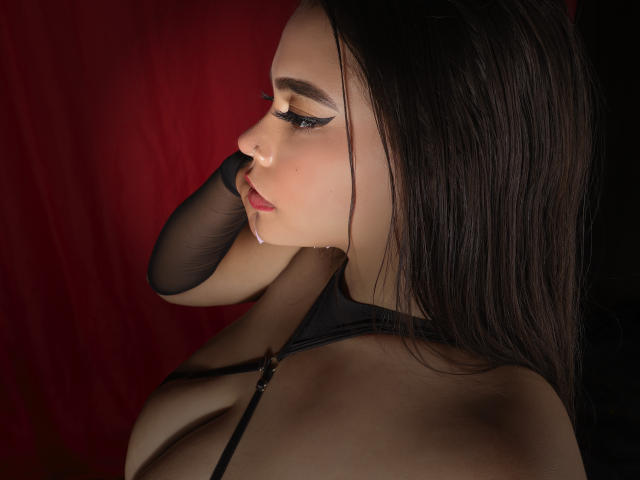 MelissaGeorgy with dark hair, black mesh arm, and drooling, looking to the side, wearing black straps, red background, and eyeliner. Her tits are partially visible.