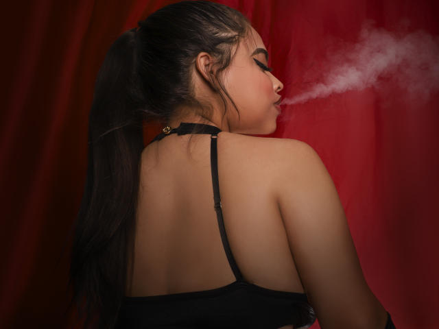 MelissaGeory with long black hair in a ponytail, black halter top, blowing white smoke from her mouth, red background, bare shoulders, looking to the right.