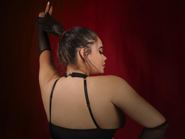 MelissaGeory with her back to the camera, arms raised, black strapless top, black gloves, and a bun, showing her ass and armpit, against a red background.