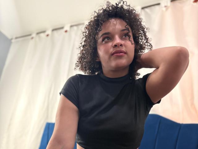 CattaleyaMolina with curly brown hair, black t-shirt, hand in hair, looking up, slightly pouty face, medium brown skin, small tits, in a softly lit room.