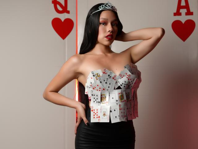 RoseSexAndFun, avec des cheveux noirs, une couronne, et un soutien-gorge rembourré de cartes, une jupe noire, une main sur la hanche, du rouge à lèvres, une pose confiante, une peau pâle, debout, sexy, des cartes recouvrant ses seins..