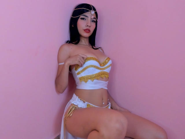 NoemyAshe with dark hair, wearing a white and gold strapless top, white and gold bottom, and a headpiece, sitting with hands on tits, pink background.
