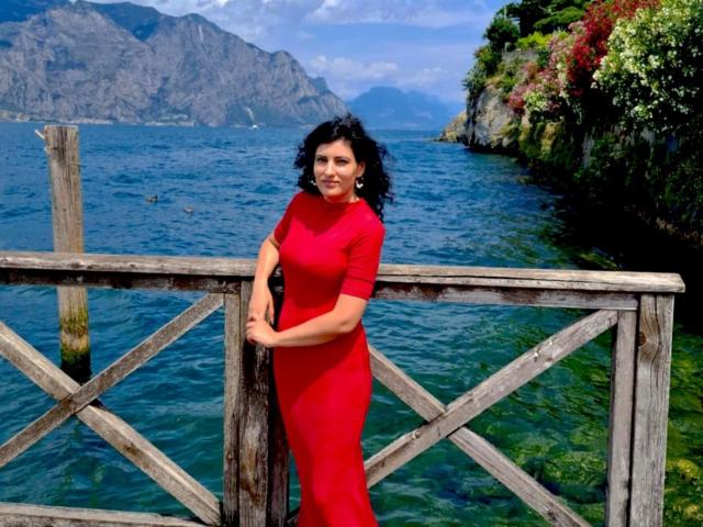 AddictedToUTS vestindo um vestido vermelho justo, segurando um corrimão de madeira, com cabelo preto ondulado, em frente a uma água azul cristalina, com vista para montanhas e vegetação exuberante ao fundo..