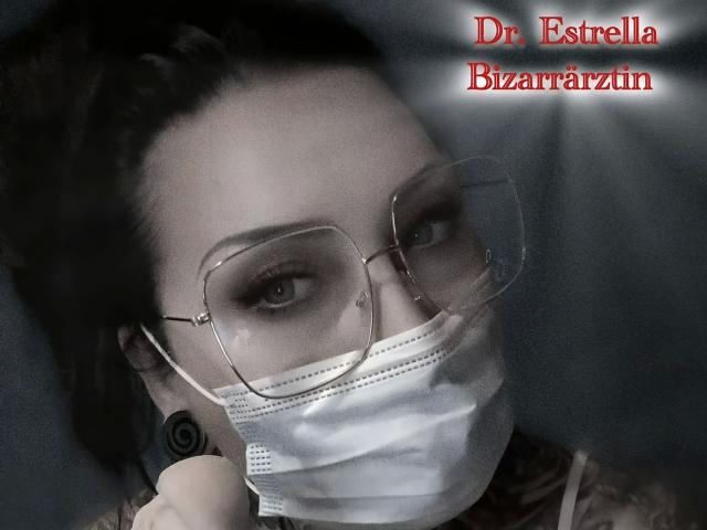StrengeTante-hot with glasses, white mask, dark hair, and black ear gauges, showing off a tattooed shoulder. Her eyes are slightly squinted. Textured, gritty photo.