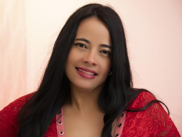PaolaValencia has long black hair, a cute smile, and is wearing a red lace top with pink buttons. She's got medium brown skin and looks sexy.