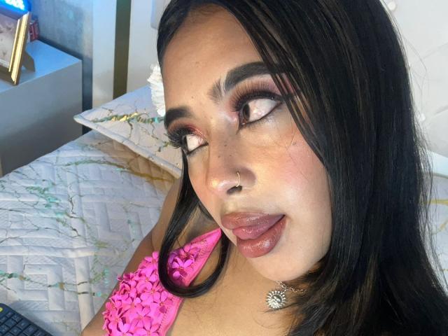 AriaMystic with dark hair, pink top, nose ring, and choker, sticking out her tongue, looking sideways with heavy makeup, including bold eyeliner and pink eyeshadow.