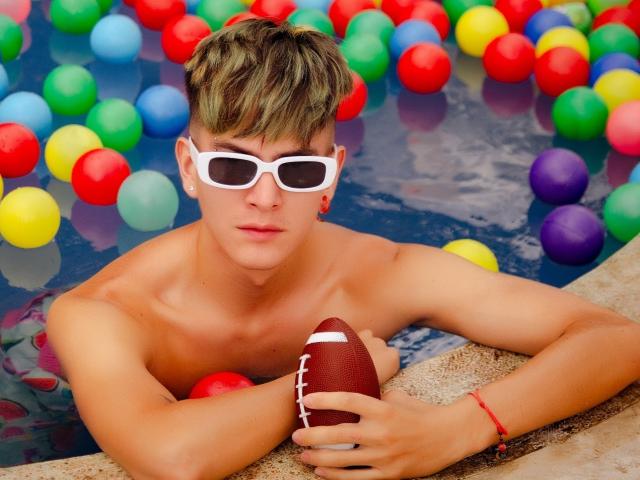 Archangel69, avec ses cheveux blonds, ses lunettes de soleil blanches et son corps bronzé, musclé et torse nu, tenant un ballon de football marron, appuyé sur le bord d'une piscine, entouré de ballons colorés..
