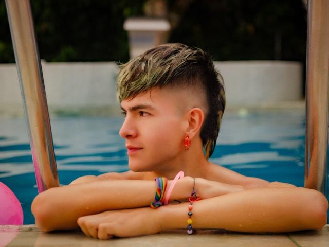 Archangel69, zonder shirt, met kort, blond gestreken haar, rode oorbellen, kleurrijke armbanden en een roze polsband, leunt op de zwemplank en kijkt opzij met een ontspannen uitdrukking..