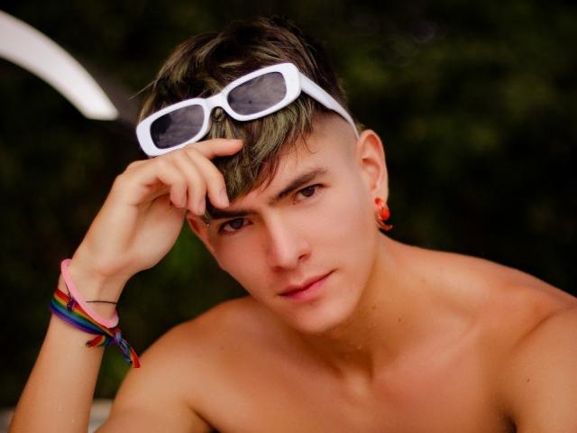 Archangel69 with short green-tinted hair, white sunglasses on head, red earrings, colorful bracelet, shirtless, looking at camera, hand on forehead, green blurry background.