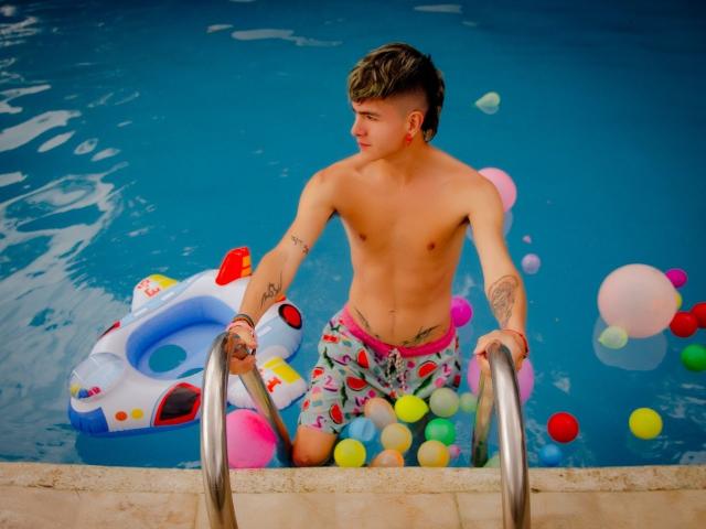 Archangel69, ohne Hemd, mit Tattoos, in bunten Badehosen, klettert aus einem blauen Pool, hält Metallstufen, umgeben von aufblasbaren Bällen und einem weißen aufblasbaren Ring..